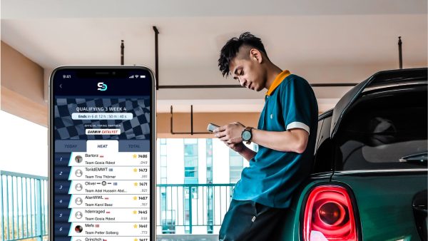 Man holding his phone and leaning against a car. iPhone showing the FIA SDC Challenge leaderboard